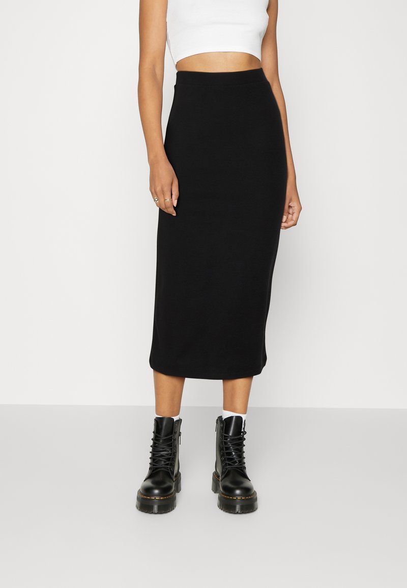 Femme portant une jupe crayon midi noire, un top court blanc, des chaussettes blanches et des bottines noires épaisses à lacets, debout devant un fond uni.