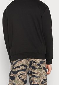 Sudadera negra de corte relajado con puños acanalados, combinada con pantalones cargo de camuflaje que presentan un patrón ondulado y multicolor.