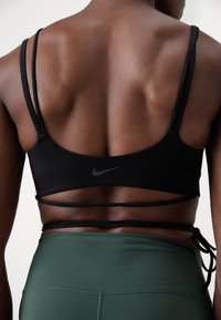 Juodas sportinis liemenukas su keliomis juostelėmis ir subtiliu Nike logotipu, derinamas su prigludusiomis tamsiai žaliomis tamprėmis. Lygi tekstūra ir minimalus dizainas.
