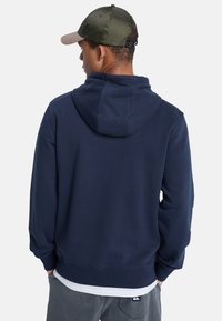 Sudadera de algodón azul marino con capucha, puños y dobladillo acanalados. Lleva puesta una camiseta blanca debajo. La vista trasera muestra un diseño mínimo y detalles de costura.