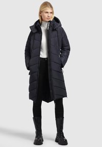 Femeie blondă îmbrăcată într-o haină lungă neagră căptușită, pullover alb cu guler înalt, pantaloni negri și cizme negre cu șireturi, stând cu mâinile în buzunare.