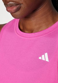 Rosa Sporttanktop aus glattem Stoff, mit rundem Ausschnitt und einem weißen Logo auf der Brust. Nahaufnahme des Ausschnitts und der Stoffstruktur.