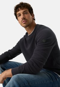 Pull-over texturé bleu marine avec un col rond et des poignets côtelés. Doté d'un patch logo noir sur la manche gauche. Associé à un jean bleu clair.