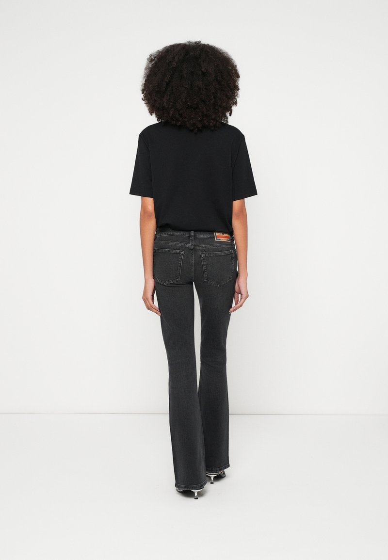Personne aux cheveux bouclés de dos, portant un t-shirt noir, un jean évasé foncé et des talons sur un fond blanc uni.