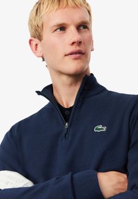Pull-over mi-zip en navy en tissu doux, avec une fermeture éclair, un col côtelé et une manche blanche avec des perforations. Détail du logo vert.