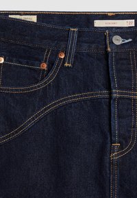 Jupe en denim bleu foncé avec poche avant visible, rivets en cuivre, passants pour ceinture et bouton métallique marqué Icon Skirt taille 27.
