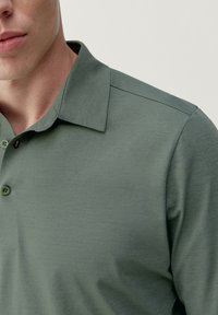 Polo shirt vert à manches courtes fabriqué en tissu texturé, avec un col et trois boutons présentant des variations de couleur subtiles.
