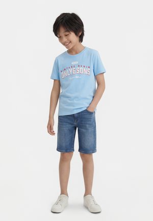 Niño sonriente con camiseta gráfica azul claro, pantalones cortos de mezclilla y zapatillas blancas, de pie con una mano en el bolsillo contra un fondo blanco.