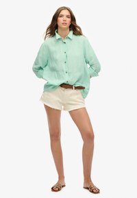 Lichtgroen linnen shirt met knoopsluiting, gecombineerd met beige versleten shorts. Een bruine riem en sandalen met ronde accenten maken de look compleet.
