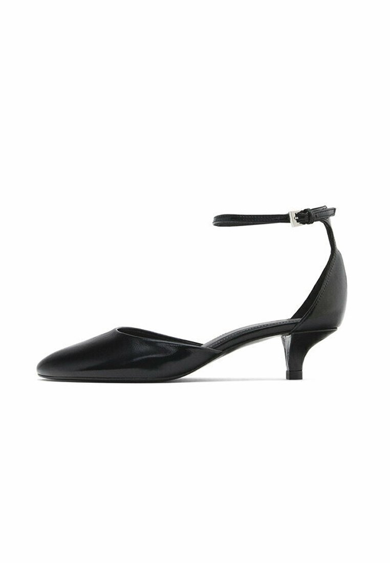 Chaussure en cuir verni noir avec un bout pointu, un petit talon compensé et une sangle ajustable à la cheville pour un maintien sécurisé. Texture lisse et design élégant.
