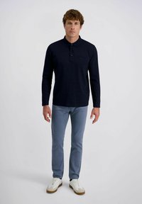 Marineblaues Langarm-Polohemd mit dezentem Streifenmuster, einzelner Brusttasche; kombiniert mit hellblauen Hosen und weißen Sneakers.