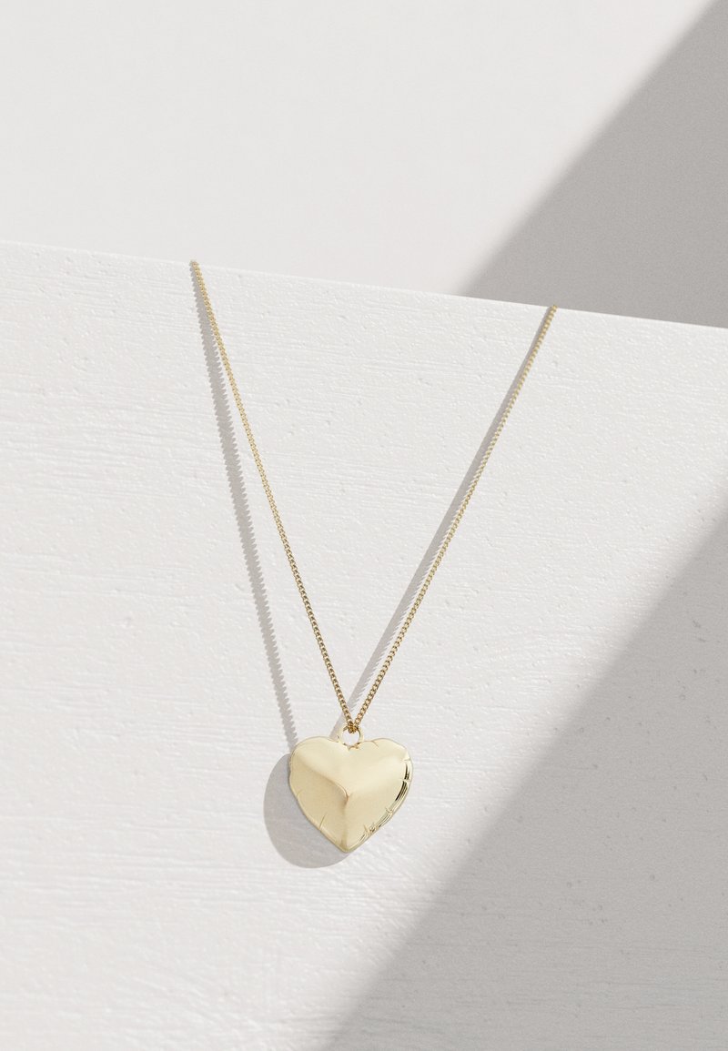 Χρυσό κολιέ με pendant σε σχήμα καρδιάς σε λεπτή αλυσίδα, γυαλιστερή επιφάνεια, ομαλή υφή, με διακριτικές αντανακλάσεις σε φωτεινό φόντο.