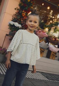 Blouse blanche avec motif de broderie florale, manches courtes à volants et ourlet peplum, associée à un jean bleu foncé. Arrière-plan de boutique de fleurs.