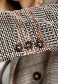 Manche de blazer à carreaux marron et beige avec trois boutons transparents, présentant une couture d'accent rouge sur le poignet. Tissu texturé.