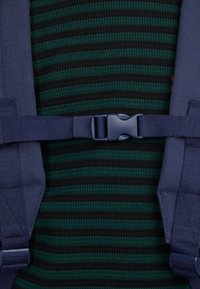 Námořnické popruhy na batoh s plastovou přezkou upevněné na zeleném a černém pruhovaném oděvu. Na popruzích je viditelná strukturovaná látka.