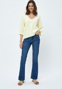 Maglione giallo a V con maniche a tre quarti, abbinato a jeans a zampa d'elefante blu e sandali beige, su uno sfondo semplice.