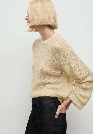 Suéter de punto en amarillo claro con un patrón de punto texturizado y abierto. Presenta mangas largas y un cuello redondo, combinado con jeans oscuros.