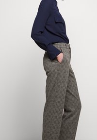 Osoba ve tmavě modré košili s dlouhým rukávem a předními kapsami, v šedých kostkovaných širokých kalhotách, stojící s jednou rukou v kapse na jednobarevném pozadí.