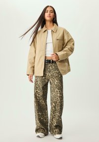 Beige oversized jakke med krave og lynlås-lomme, parret med brede olivengrønne leopardmønstrede bukser og hvide sneakers.