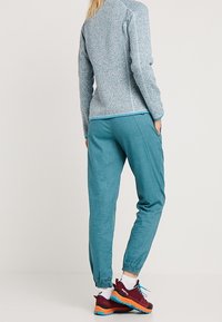 Vrouw in een lichtblauwe fleecejas, met teal joggingbroek en bordeauxoranje sportschoenen, staande met handen in haar zakken, met haar rug naar de camera gekeerd.