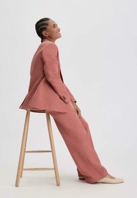 Lichtroze op maat gemaakte pak met een fitted blazer en wijde broekspijpen, gemaakt van gladde stof, gecombineerd met beige instappers, zittend op een houten kruk.