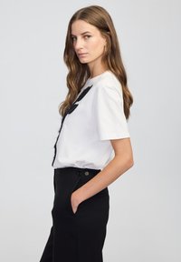 Vrouw met lang bruin haar, gekleed in een witte shirt met korte mouwen en zwart ontwerp, en zwarte high-waisted broek, staand in profiel met één hand in de zak.