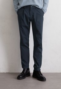 Donkerblauwe op maat gemaakte broeken met een gladde textuur, voorzien van een knoopsluiting en plooien. GDragen met zwarte leren veterschoenen.