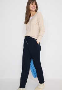 Femme souriante portant un pull beige, un pantalon noir à cordon de serrage, des baskets blanches, tenant une veste bleue, debout devant un fond blanc.