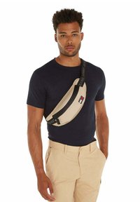 Bolso de cintura beige con una correa negra, que presenta un parche con el logo, que se lleva cruzado sobre el pecho con una camisa azul marino y pantalones caquis. Textura de tela suave.