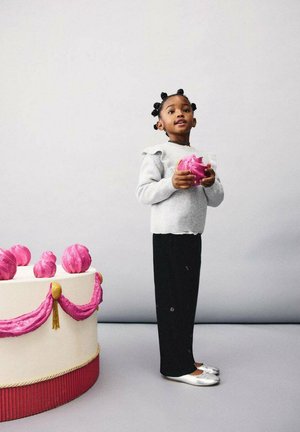 Jeune fille en sweat gris et pantalon noir tenant un grand macaron rose près d'une table de gâteaux décorée avec des détails en glaçage rose.
