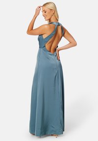 Dlouhá saténová šatna v jemně modrém odstínu. Má hluboký výstřih na zádech, široké ramínka a splývavý střih. Hladká textura a minimalistický design.