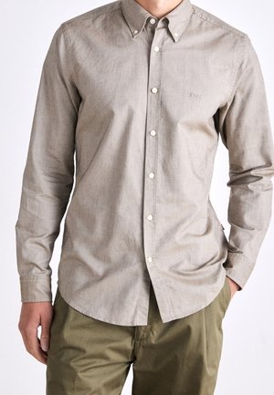 Mand iført en lys beige skjorte med knapper og lange ærmer med "BOSS"-logo på brystet, sat sammen med olivengrønne bukser, står med den ene hånd i lommen.