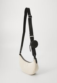 Beige fabric shoulder bag with a curved shape, black leather accents, an adjustable strap, and a round zippered pouch attached.