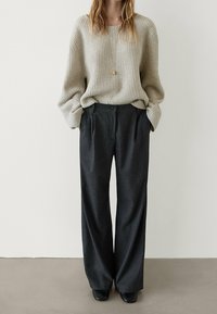 Pull beige à côtes avec des manches longues et une coupe décontractée, associé à un pantalon gris foncé à rayures, avec un devant plat et des poches.