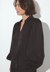 Black satin blouse featuring a V-neck, buttons down the front, and puffed long sleeves with elastic cuffs. Smooth texture and elegant design.