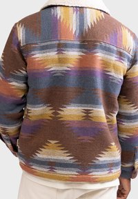 Veste en polaire à motifs marron avec un col en fausse fourrure, présentant des motifs géométriques en bleu, jaune et violet. Coupe régulière avec des détails à boutons.