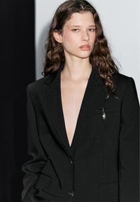 Mladý člověk s dlouhými vlnitými vlasy má na sobě černý sako s hlubokým výstřihem do V a stříbrnou broží, stojí proti černo-bílému pozadí.