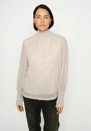 Light beige long-sleeve blouse with a high ruffled neckline and elasticized cuffs. Sheer, textured fabric overlaid. Worn with dark jeans.