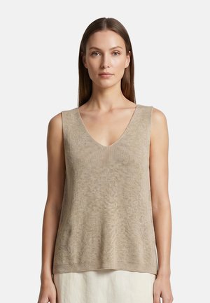 Donna con capelli lisci che indossa un top beige in maglia senza maniche con scollo a V e una gonna chiara su uno sfondo semplice.