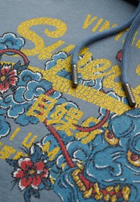 Sudadera azul con un gráfico amarillo agrietado que presenta un dragón azul y patrones florales. Cuerdas de ajuste azul claro y detalles de hardware plateados.