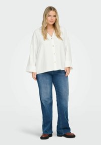 Camicia bianca con colletto a V e maniche lunghe e morbide, abbinata a jeans a zampa d'elefante blu e stivali marroni.