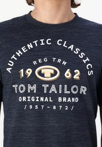 Tums zilgans kokvilnas T-krekls ar apaļu kaklu, kurā ir balti un zeltaini izdrukāti teksti: "AUTENTISKIE KLASIKAS 1962 TOM TAILOR ORIĢINĀLĀ ZĪMOLS."