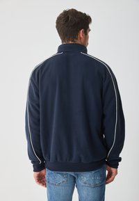 Sudadera de forro polar navy con cuello alto, con ribetes blancos en los hombros y dobladillo acanalado. Se lleva con pantalones vaqueros azules, vista desde atrás.