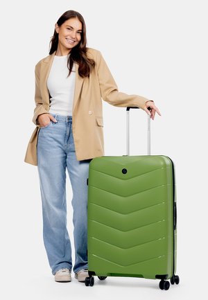 Grüne Hartschale-Koffer mit einem Chevron-Muster, multidirektionale Räder und einem ausziehbaren Griff, neben einer Person in Freizeitkleidung platziert.