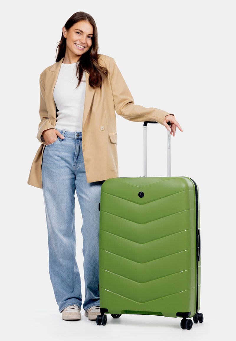 Zelený tvrdý kufr s vzorem chevron, s kolečky pro více směrů a teleskopickou rukojetí, stojící vedle osoby v neformálním oblečení.