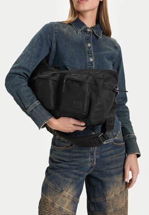 Mujer con chaqueta vaquera abotonada y jeans sostiene una gran riñonera negra con varios bolsillos con cremallera y una correa ajustable.