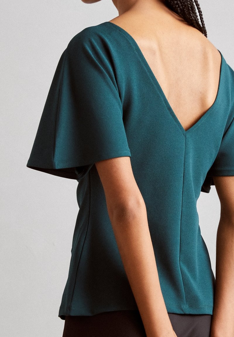 Femme portant un haut bleu sarcelle à manches courtes avec un profond décolleté en V dans le dos et des manches larges sur un fond uni.