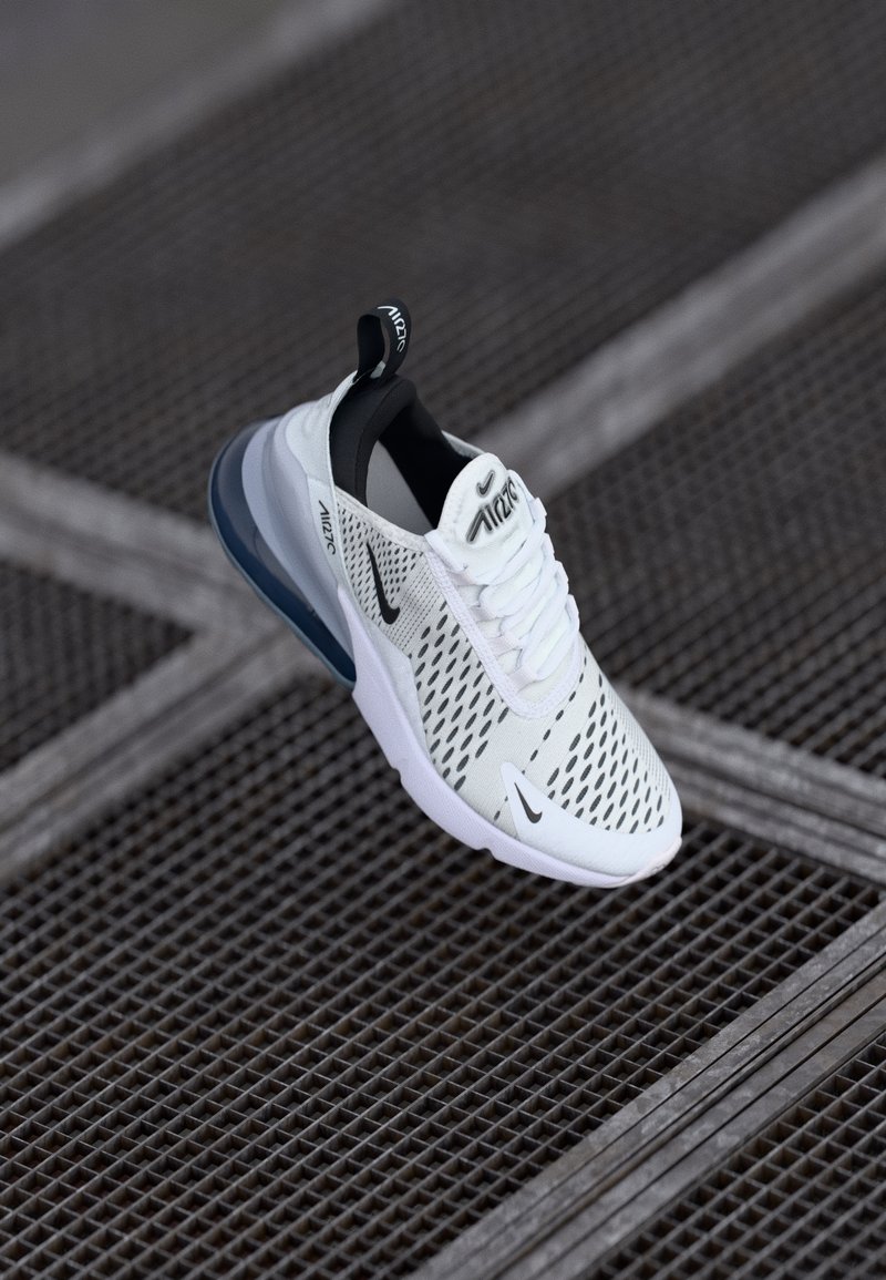 Baskets blanches Nike Air Max avec des accents noirs et un amorti à air visible, présentées au-dessus d'une surface de sol en grille métallique.