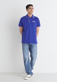 Modrá polo tričko s golierom a bielymi detailmi, skombinované so svetlomodrými džínsami a bielymi teniskami. Voľný strih s krátkymi rukávmi a detailom loga.