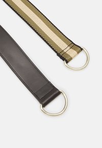 Due estremità di cintura con anelli in metallo dorato; una in pelle nera, l'altra in tessuto intrecciato verde e beige con testo sfumato sulla striscia beige.